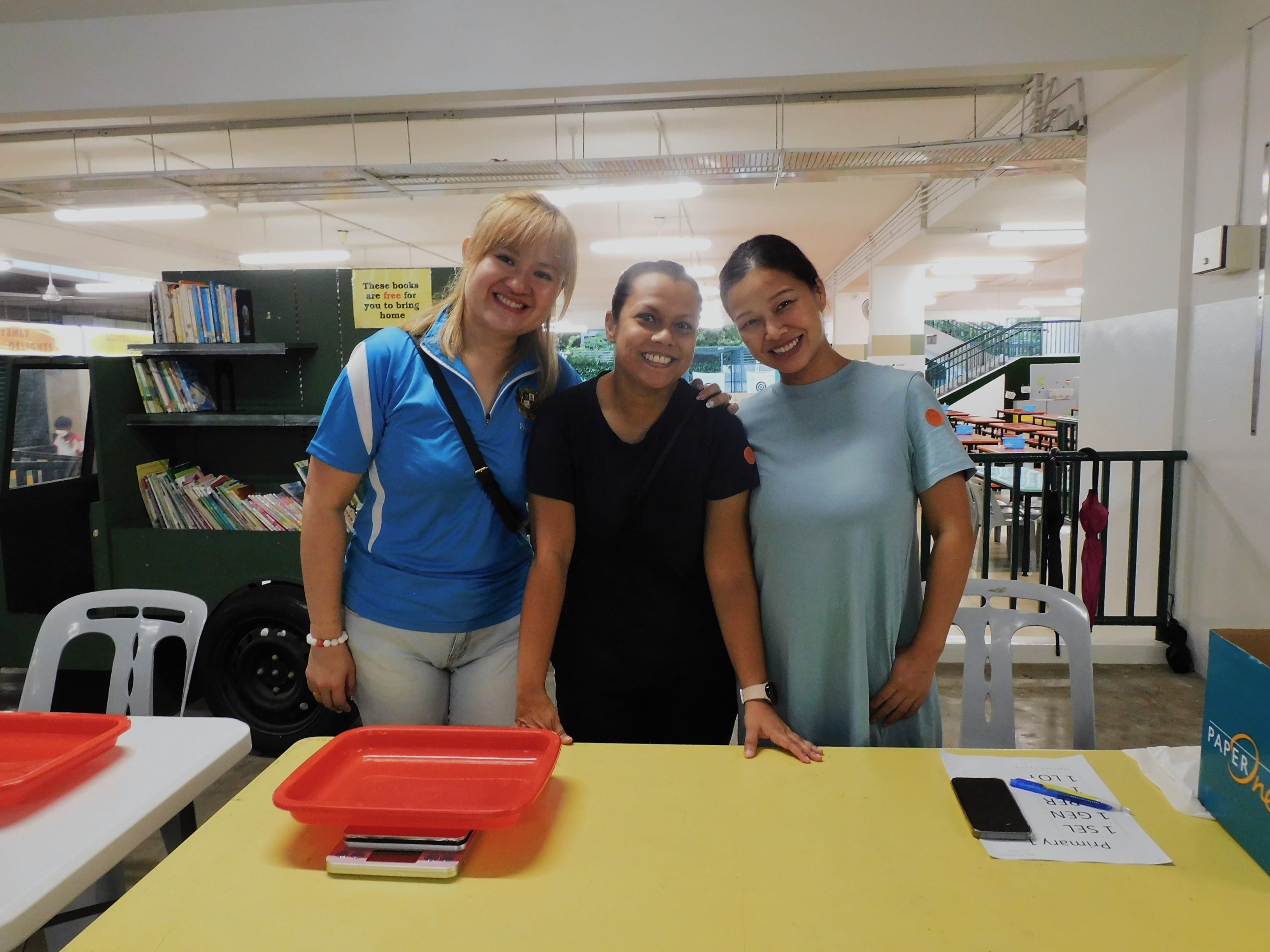 Three smiling women stand together behind a yellow table in a school canteen area with bookshelves in the background.